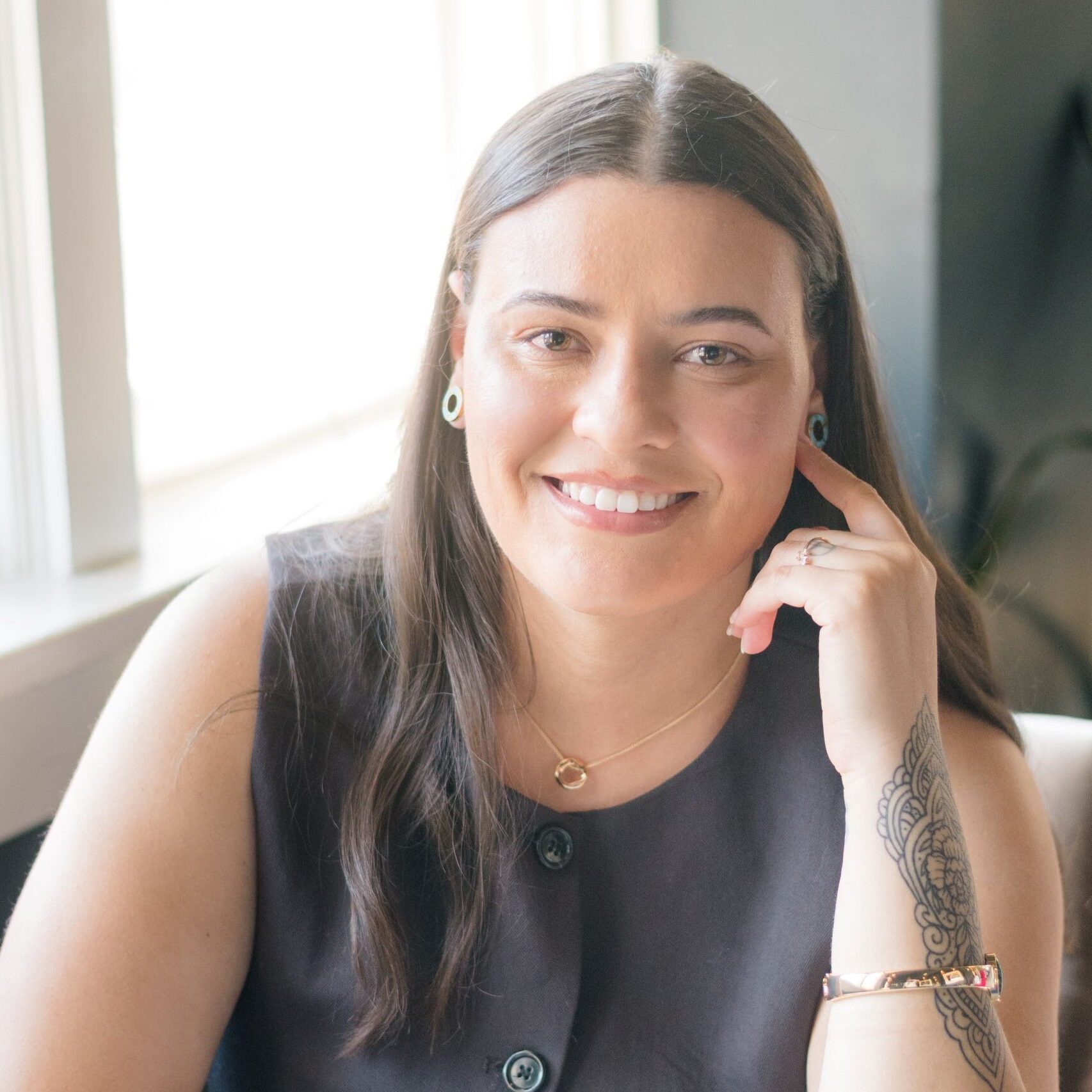 Circular casual-professional style of photo of Xavia, smiling with her teeth, one hand resting on a coffee table next to a white mug, one hand against her head. Long brown hair, tan skin, black sleeveless blouse, plant and window in the background.